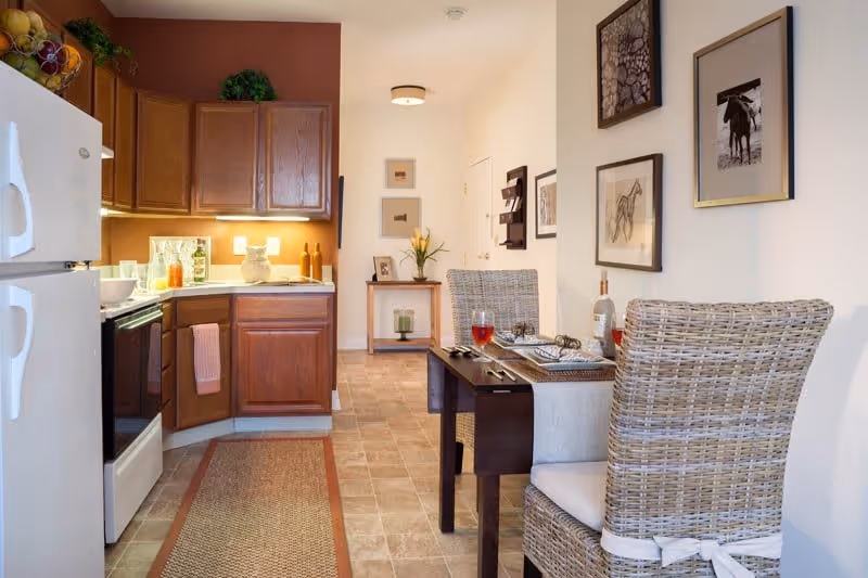 A cozy kitchen and dining area in a senior living facility. The kitchen features wooden cabinets, a white refrigerator, and a stove with a pink towel hanging on the handle. The dining area has a dark wooden table set for two with woven chairs, a bottle of wine, and glasses. The walls are decorated with framed pictures, and a small table with a vase of flowers is visible in the background.