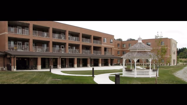 Three-story brick senior living building with balconies and a white gazebo on a landscaped lawn with winding walkways.