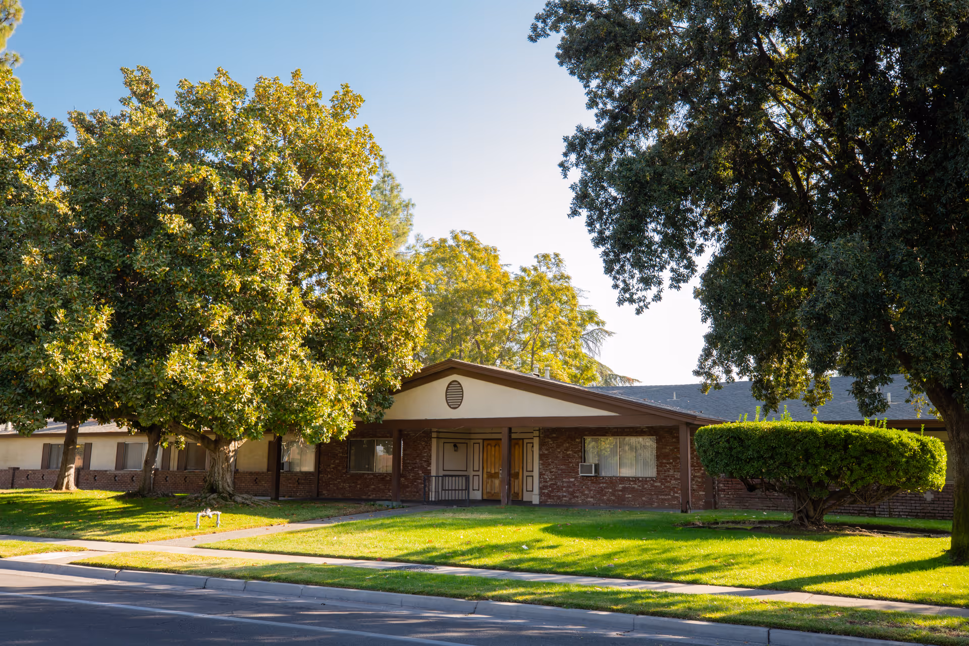 Single-story brick building with a gabled entrance, large shade trees, and a manicured lawn along the street.