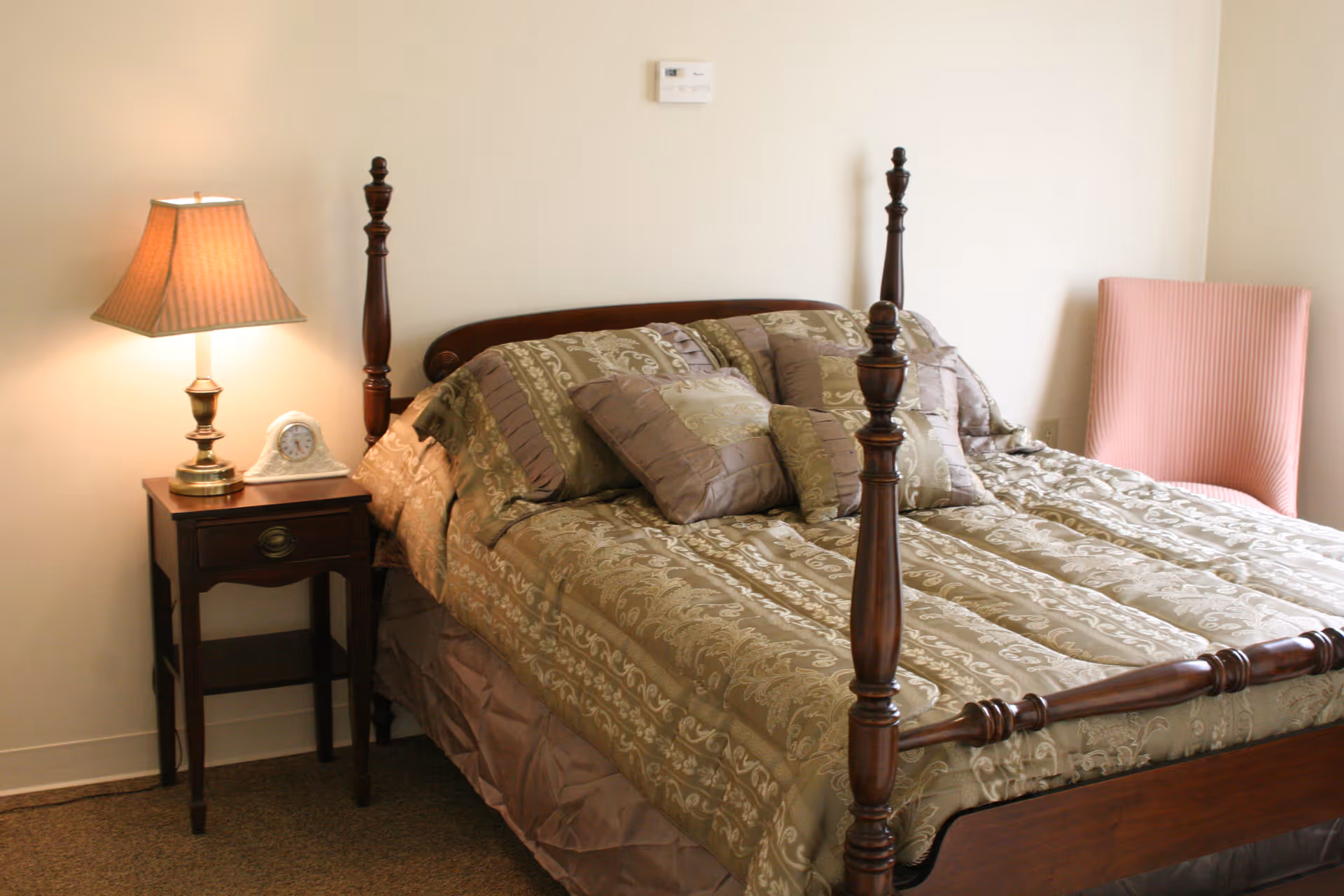 A bedroom with a wooden four-poster bed covered with a patterned green and beige comforter and several matching pillows. To the left of the bed is a wooden nightstand with a brass lamp and a small clock. A pink upholstered chair is positioned against the wall to the right of the bed.