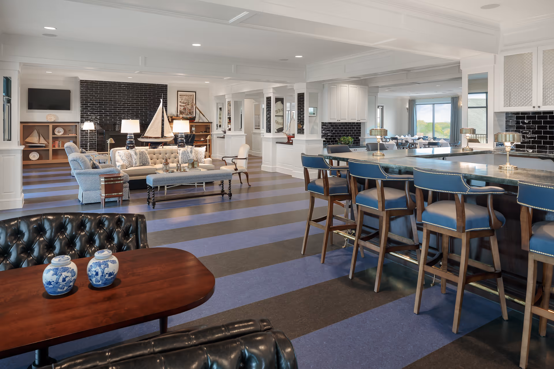 A spacious and well-lit common area with a mix of seating options including a bar with blue cushioned stools, a tufted leather chair, and a cozy sitting area with a sofa, armchairs, and a bench. The room features a striped floor, white walls with columns, a black tiled fireplace, decorative sailboat models, and large windows letting in natural light.