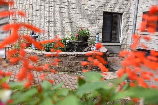 Small outdoor courtyard with a raised stone fountain and planter in front of a brick building, seen through blurred red flowers in the foreground.
