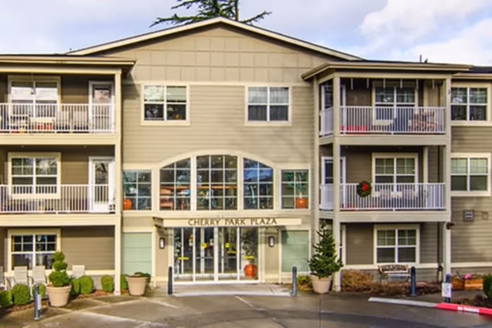 Front exterior view of Cherry Park Plaza, a three-story building with balconies, large windows, and a main entrance with glass doors. There are potted plants and small bushes near the entrance and a bench on the right side.