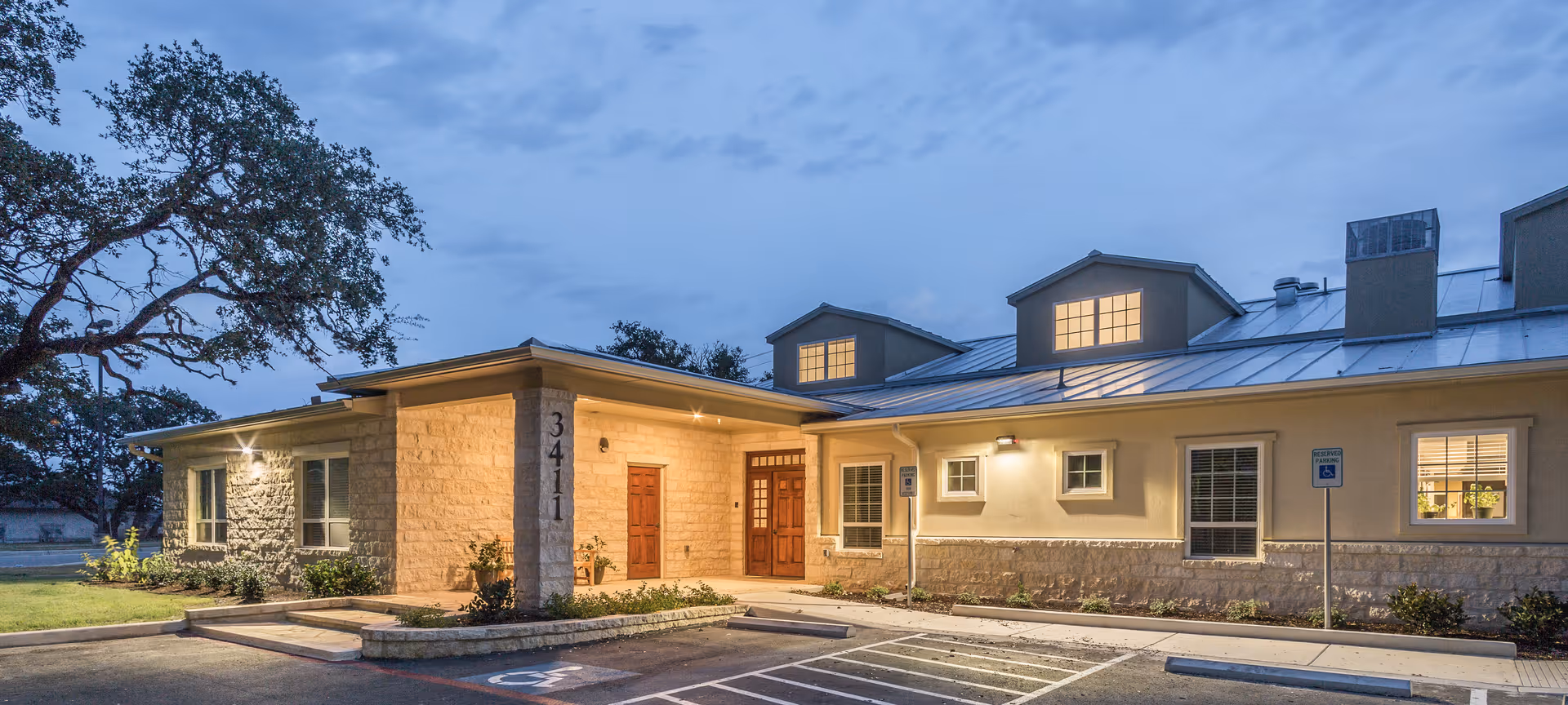 Exterior view of Bader House Memory Care of Shavano Texas building at dusk, showing a single-story structure with stone and beige walls, a metal roof with dormer windows, illuminated entrance, and a parking area with handicap parking spaces in front.