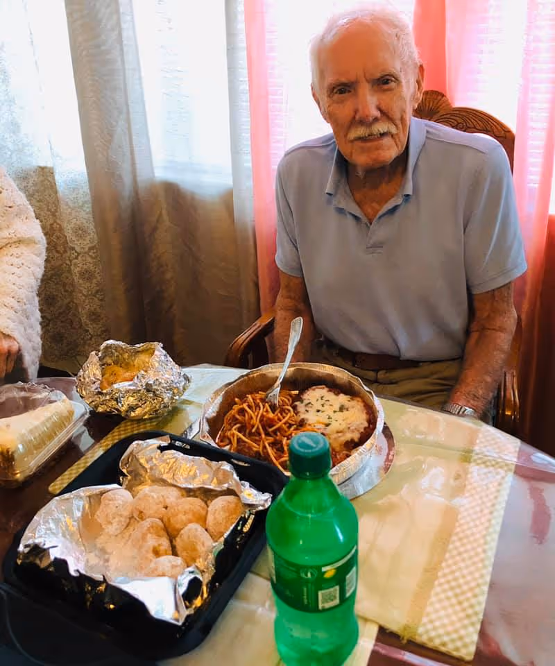An elderly man with white hair and a mustache sits at a dining table with a plate of spaghetti and baked dish, a green soda bottle, and containers of food including powdered donuts and a baked potato wrapped in foil. The background shows lace curtains and pink drapes.
