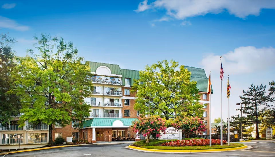 Exterior view of Bedford Court senior living community building with green roofs, balconies, and a landscaped roundabout featuring flowers and trees under a blue sky with some clouds.