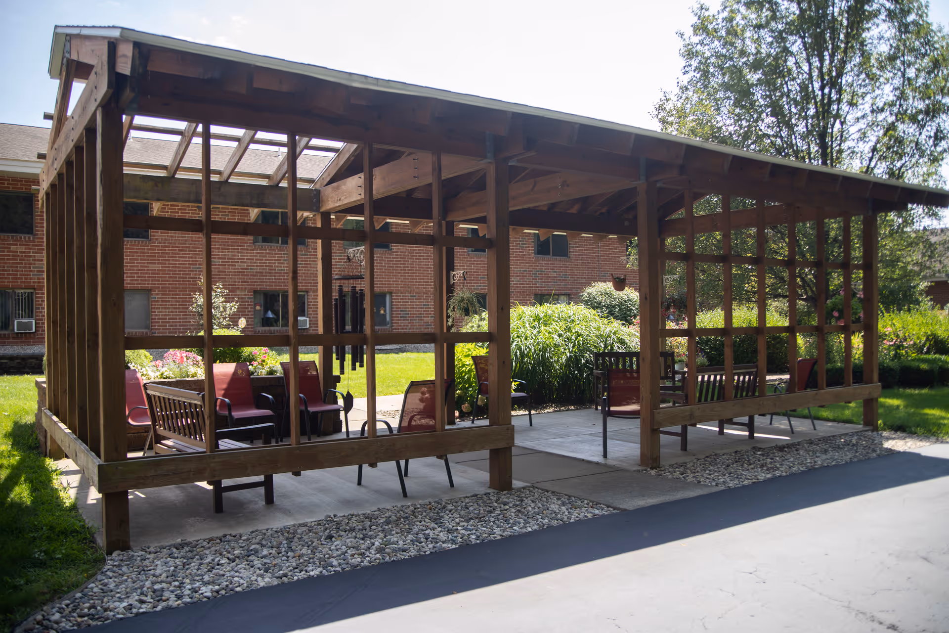 Outdoor wooden pergola structure with seating including benches and chairs, surrounded by greenery and flowers, with a brick building in the background under a clear sky.