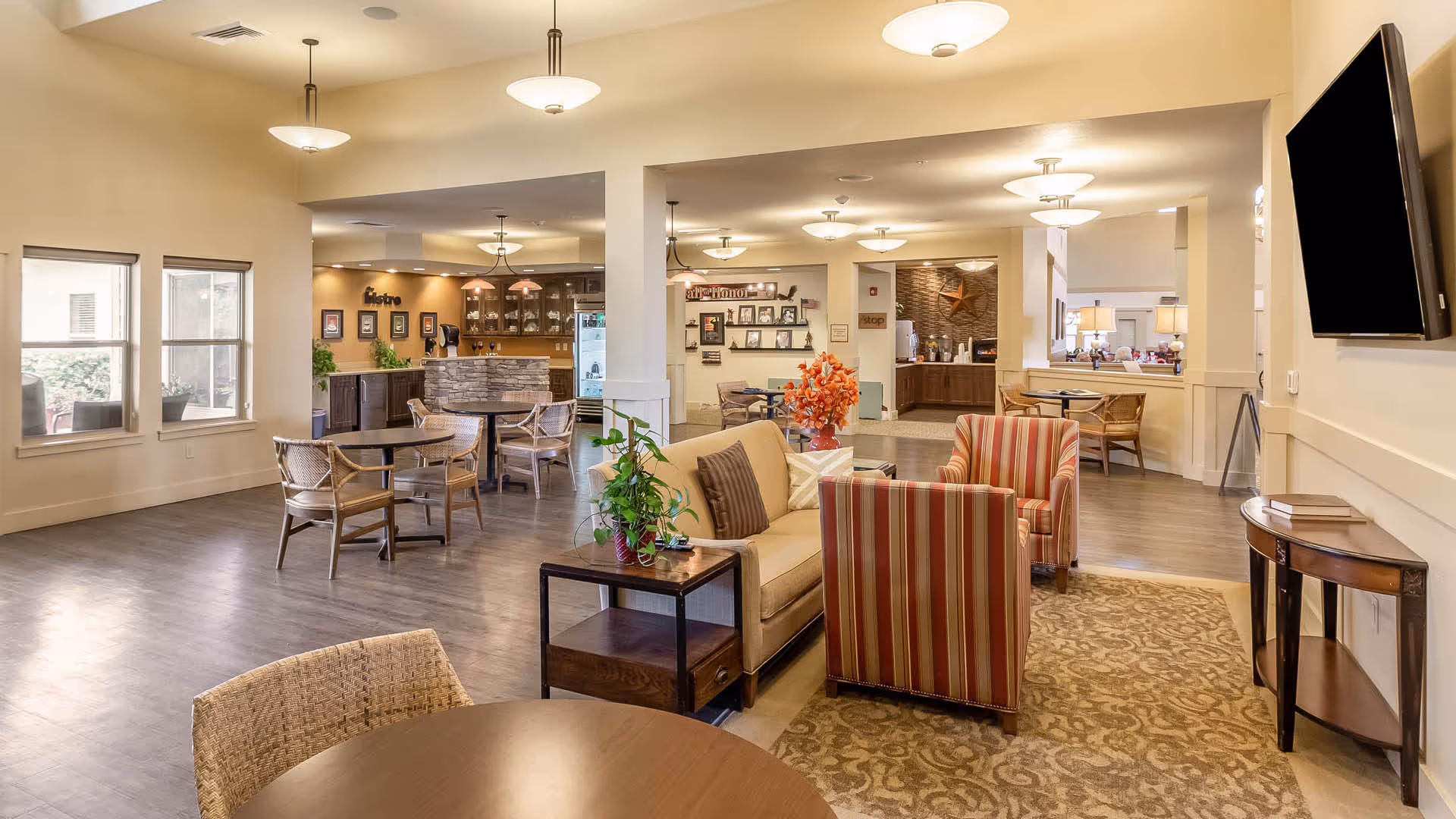 Bright, open common lounge area with sofas, armchairs, dining tables, and a wall-mounted TV.