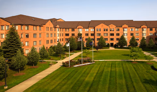 Brick multi-story senior living building surrounding a landscaped central lawn with flagpoles and walkways.