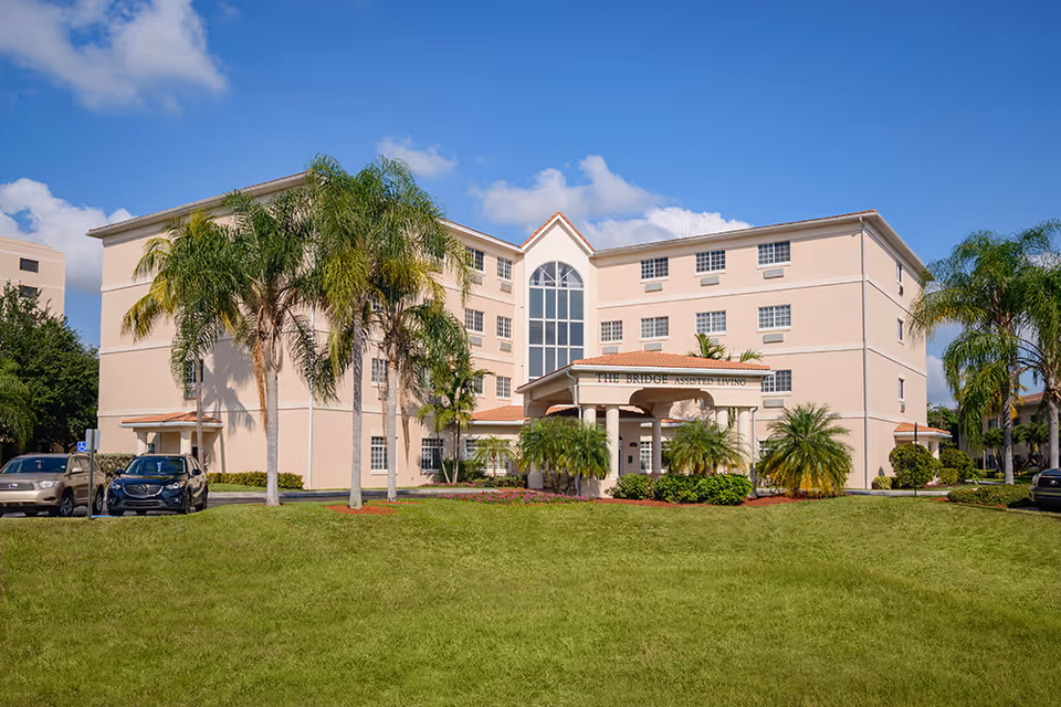 Front view of a peach-colored multi-story assisted living building with palm trees, a grassy lawn, and parked cars.
