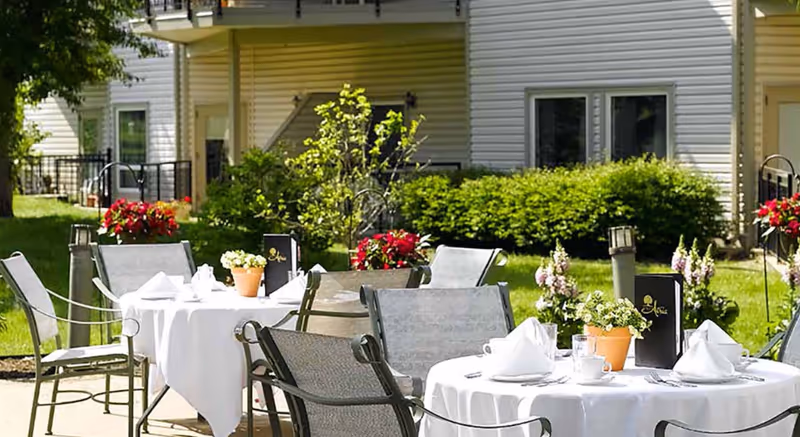 Outdoor patio with round tables set with white tablecloths and chairs in front of a residential building, surrounded by potted flowers and greenery.