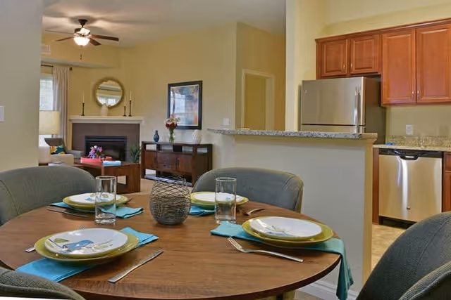 A dining area with a round wooden table set for four with plates, glasses, and turquoise napkins. Behind the dining area is a kitchen with stainless steel appliances including a refrigerator and dishwasher, wooden cabinets, and a granite countertop. In the background, there is a living room with a fireplace, a round mirror above the mantel, a wooden sideboard with decorative items, and a window with curtains.