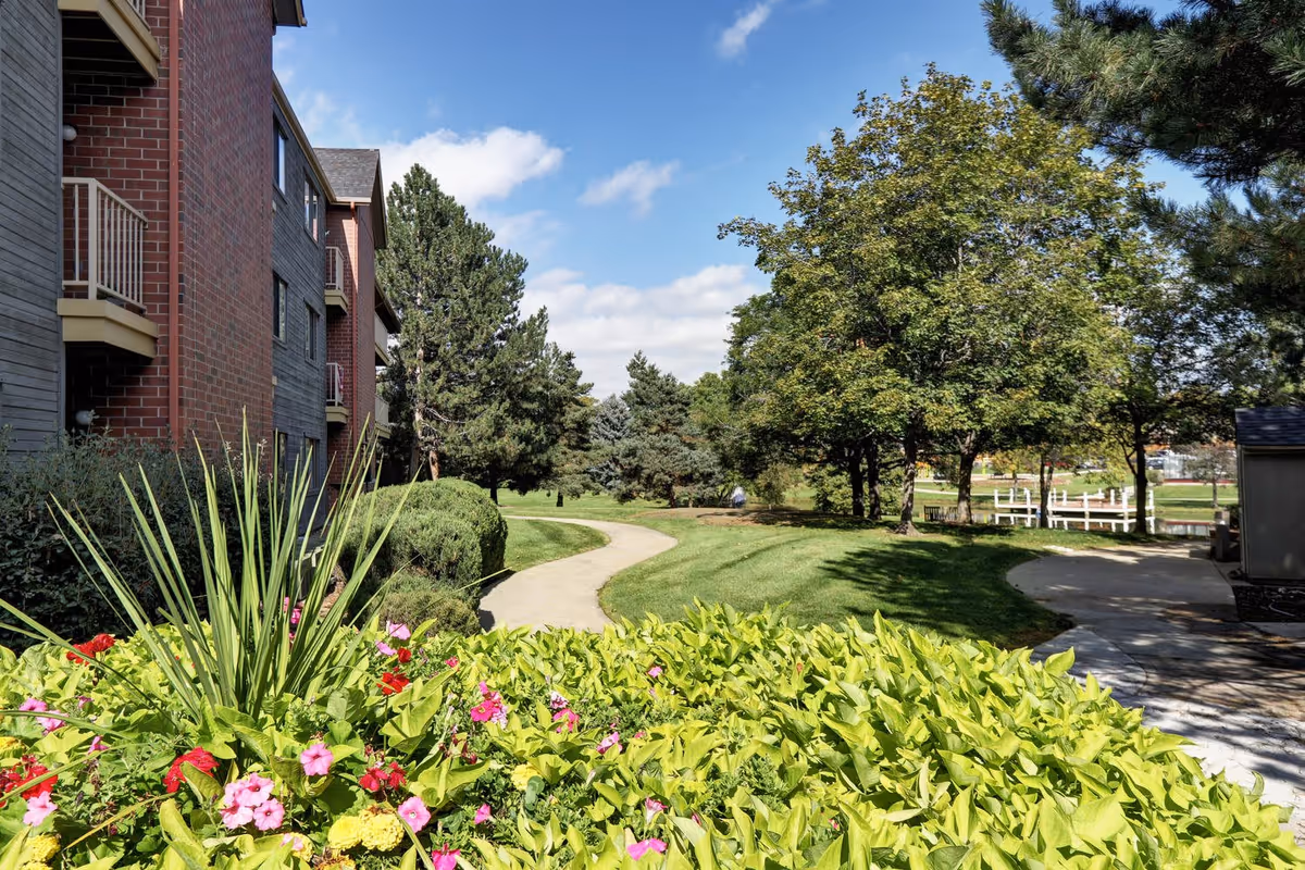 A landscaped outdoor area at a senior living facility featuring a winding concrete pathway surrounded by green grass, various trees, and colorful flowering plants. On the left side, there is a multi-story brick and wood building with balconies. The sky is blue with some clouds.