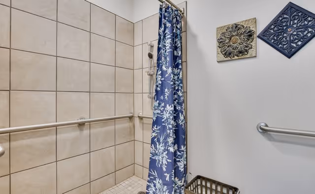 A tiled shower area with beige tiles and a blue floral shower curtain partially drawn. There are metal grab bars on the tiled walls and a handheld showerhead. On the adjacent white wall, two decorative wall hangings are visible above a metal basket.