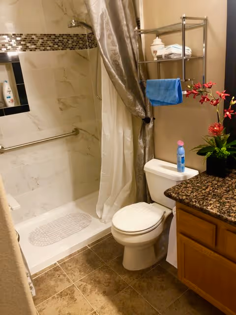 Bathroom with a walk-in shower and curtain, toilet next to a granite-topped vanity, wall shelf with towels, and a floral arrangement.