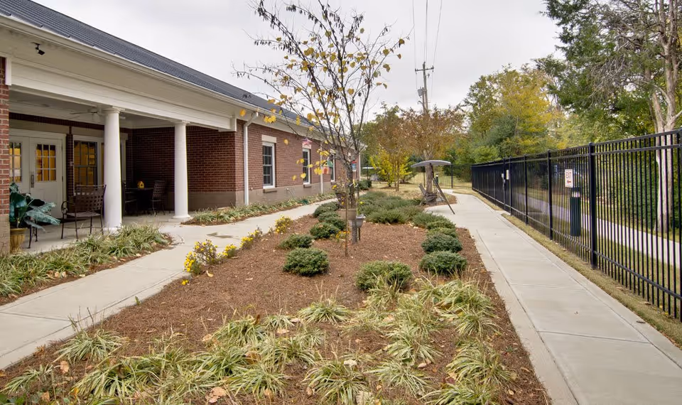 Outdoor garden area at Schilling Gardens featuring a brick building with white columns on the left, a paved walkway, landscaped plants and shrubs, a small tree in the center, and a black metal fence on the right side.