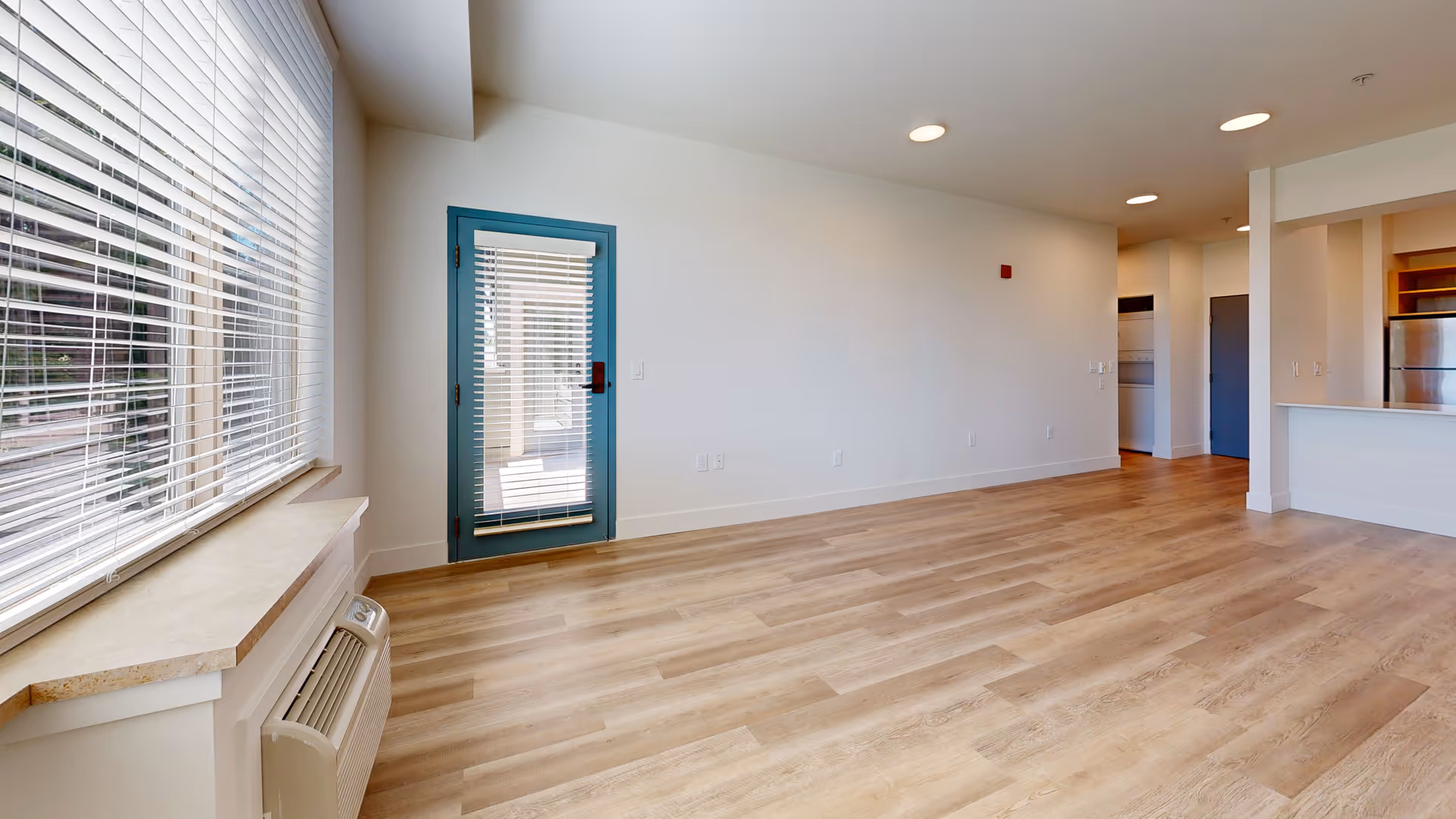 Bright open living area with wood-look flooring, a large window with blinds, a teal-framed door, and a partial view of the kitchen.