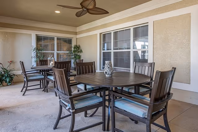 Covered outdoor patio area with round tables and cushioned chairs, ceiling fan, potted plants, and large windows on the wall.