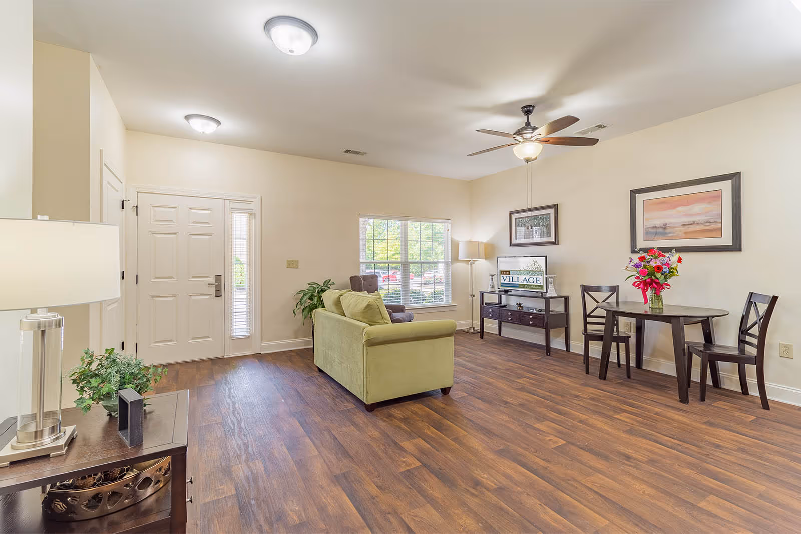 A bright and spacious living room with wood flooring, a green sofa, a small dining table with two chairs and a vase of flowers, a ceiling fan with light, framed artwork on the walls, a window with blinds, and a door with sidelights. There is also a side table with a lamp and a plant, and a console table with a lamp and a sign that reads 'The Village'.