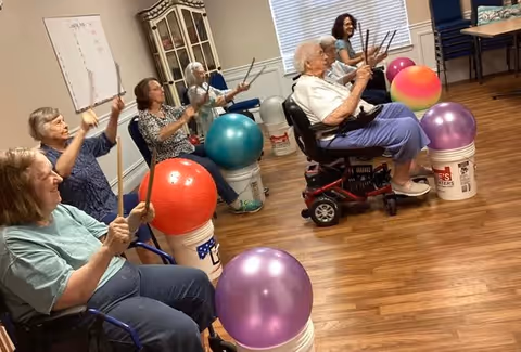 A group of elderly individuals seated in a room participating in a drumming exercise using drumsticks and colorful exercise balls placed on white buckets. The room has wooden flooring, a whiteboard on the wall, and a cabinet with glass doors.