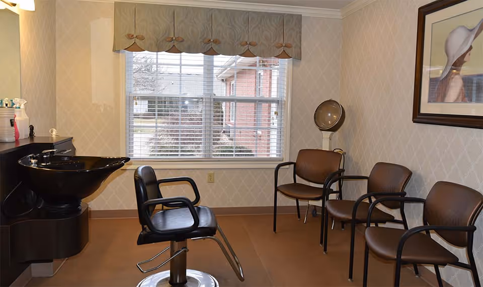 Interior of a small salon room with a black hair washing sink and chair on the left, three brown chairs lined up against the right wall, a large window with blinds and a valance, and a framed picture on the wall.