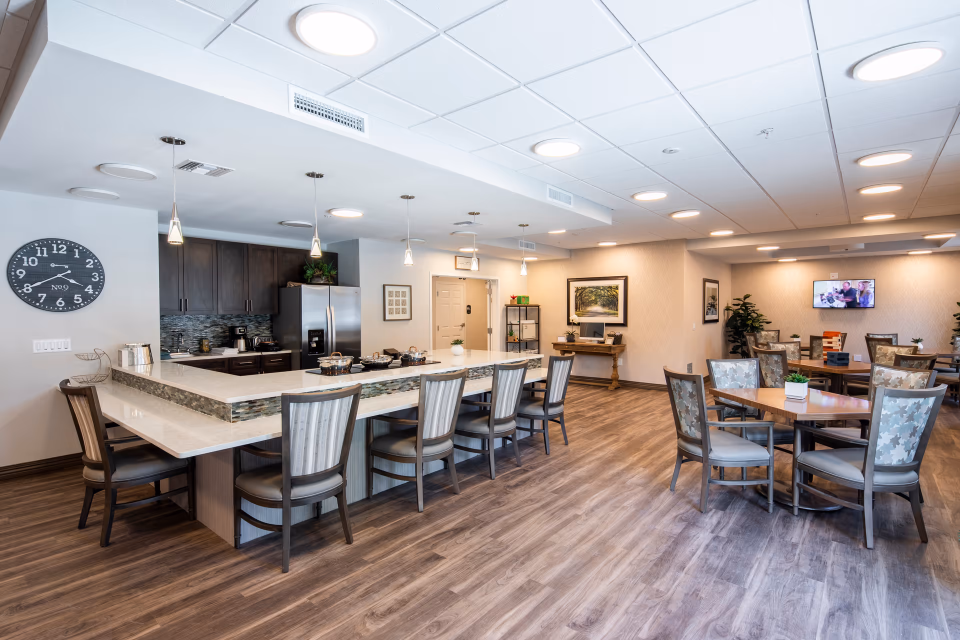 A spacious and well-lit common area in Sonata Vero Beach featuring a large kitchen island with several chairs, pendant lights hanging from the ceiling, a wall clock, and a stainless steel refrigerator. The room also includes multiple dining tables with chairs, a television mounted on the wall, framed artwork, and decorative plants, all set on wood-style flooring.