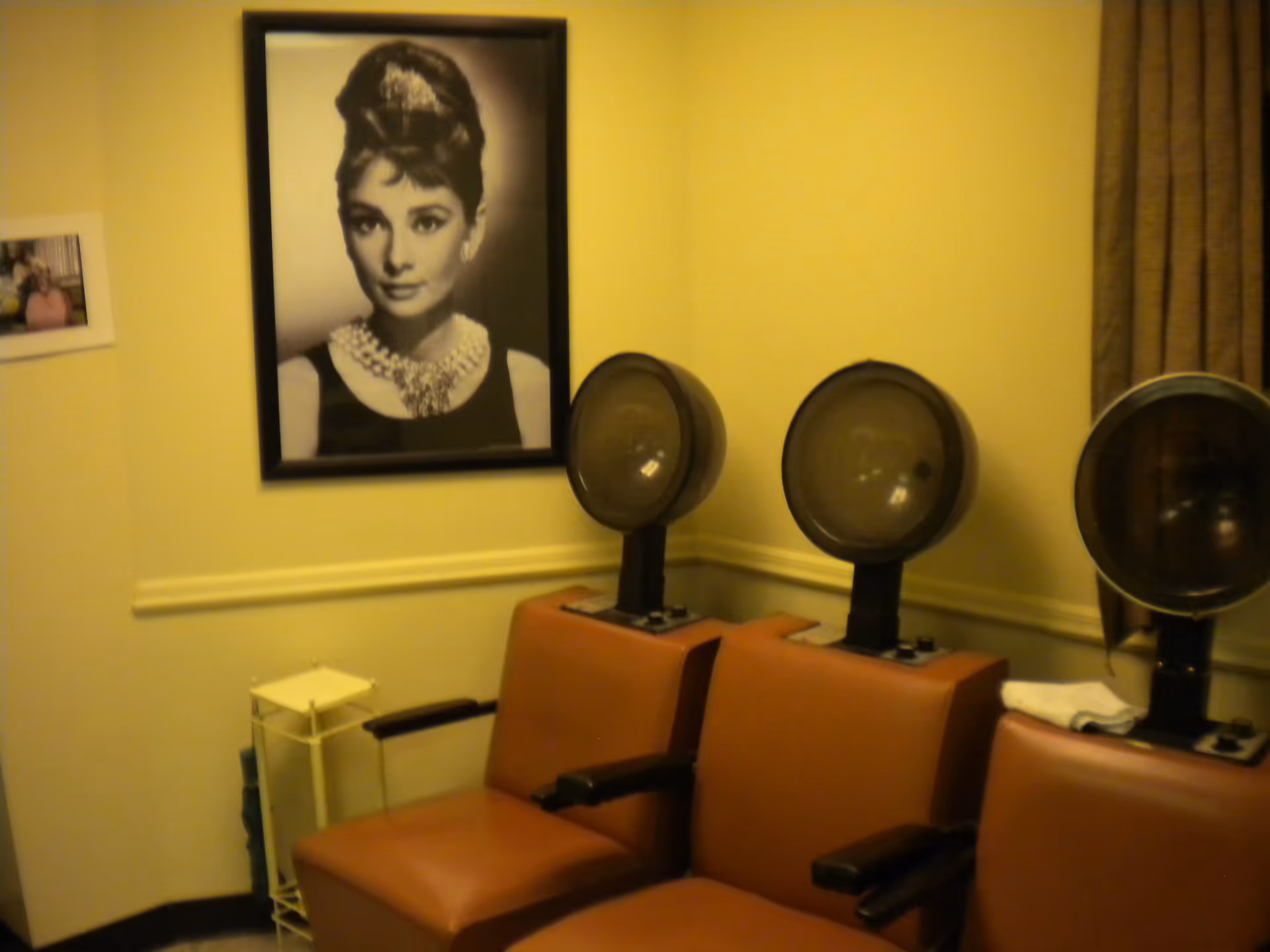 Small salon-style room with three hooded hair dryers above red salon chairs and a large framed black-and-white portrait on the wall.