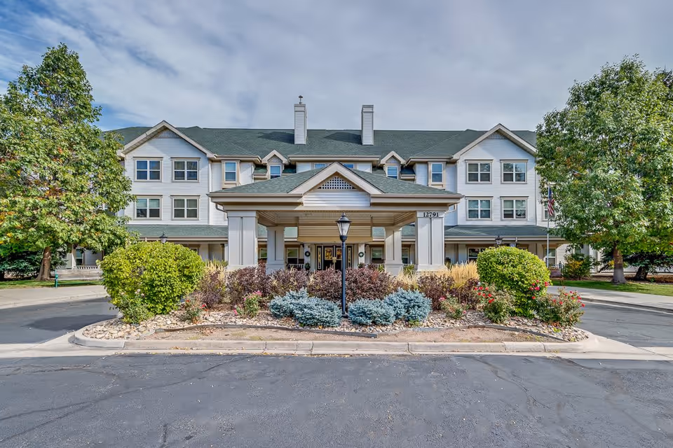 Front exterior view of Woodlake Legacy Senior Living building with a covered entrance, surrounded by landscaped bushes, trees, and a paved driveway under a partly cloudy sky.