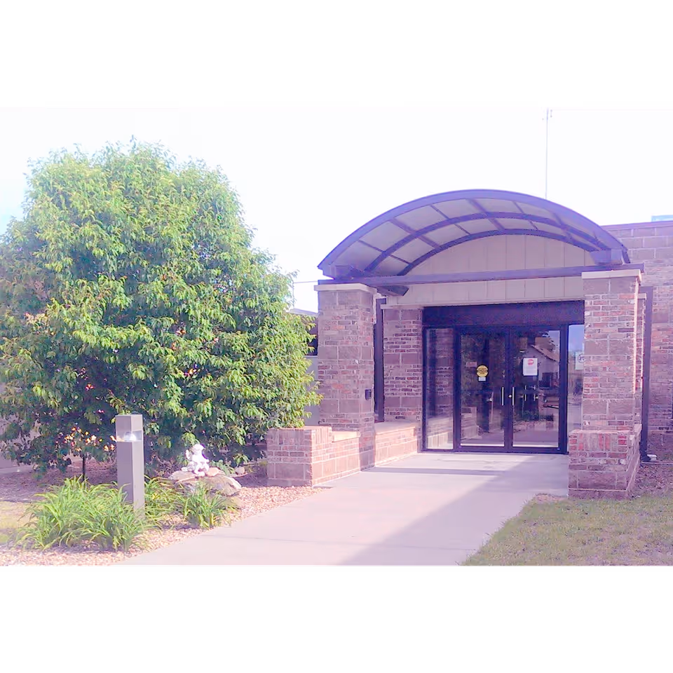 Entrance to a building with a curved metal awning above glass double doors. The building is made of brick, and there is a large leafy green tree and some plants on the left side of the walkway leading to the entrance.