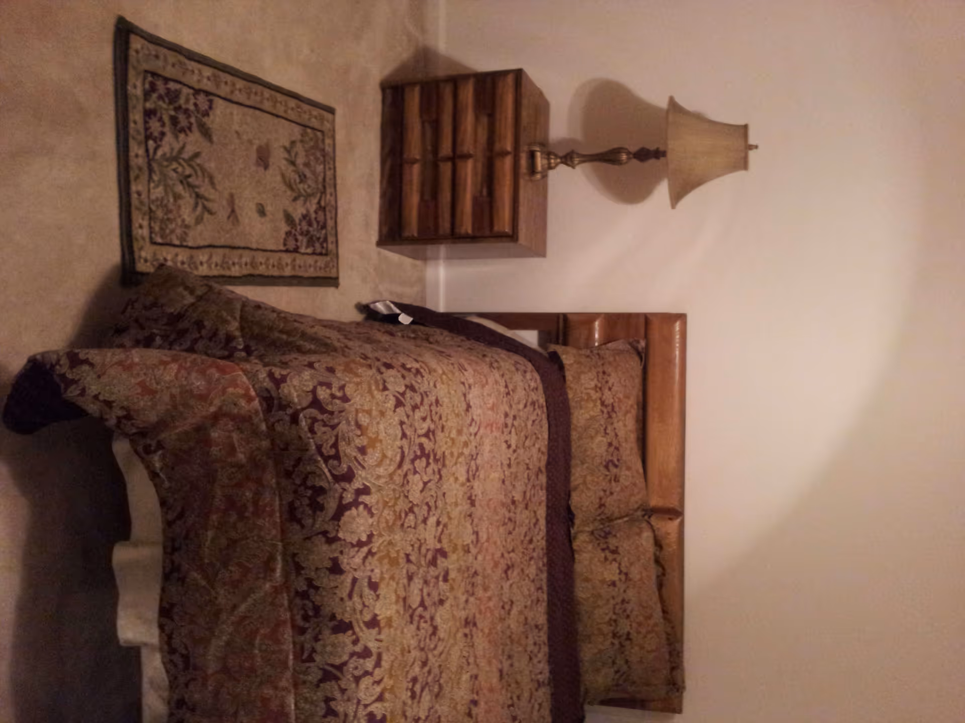 A bedroom with a bed covered in a patterned bedspread in shades of brown and gold. There is a wooden nightstand with multiple drawers next to the bed, topped with a brass lamp with a beige lampshade. A small decorative rug is placed on the carpeted floor beside the bed. The walls are plain and light-colored.