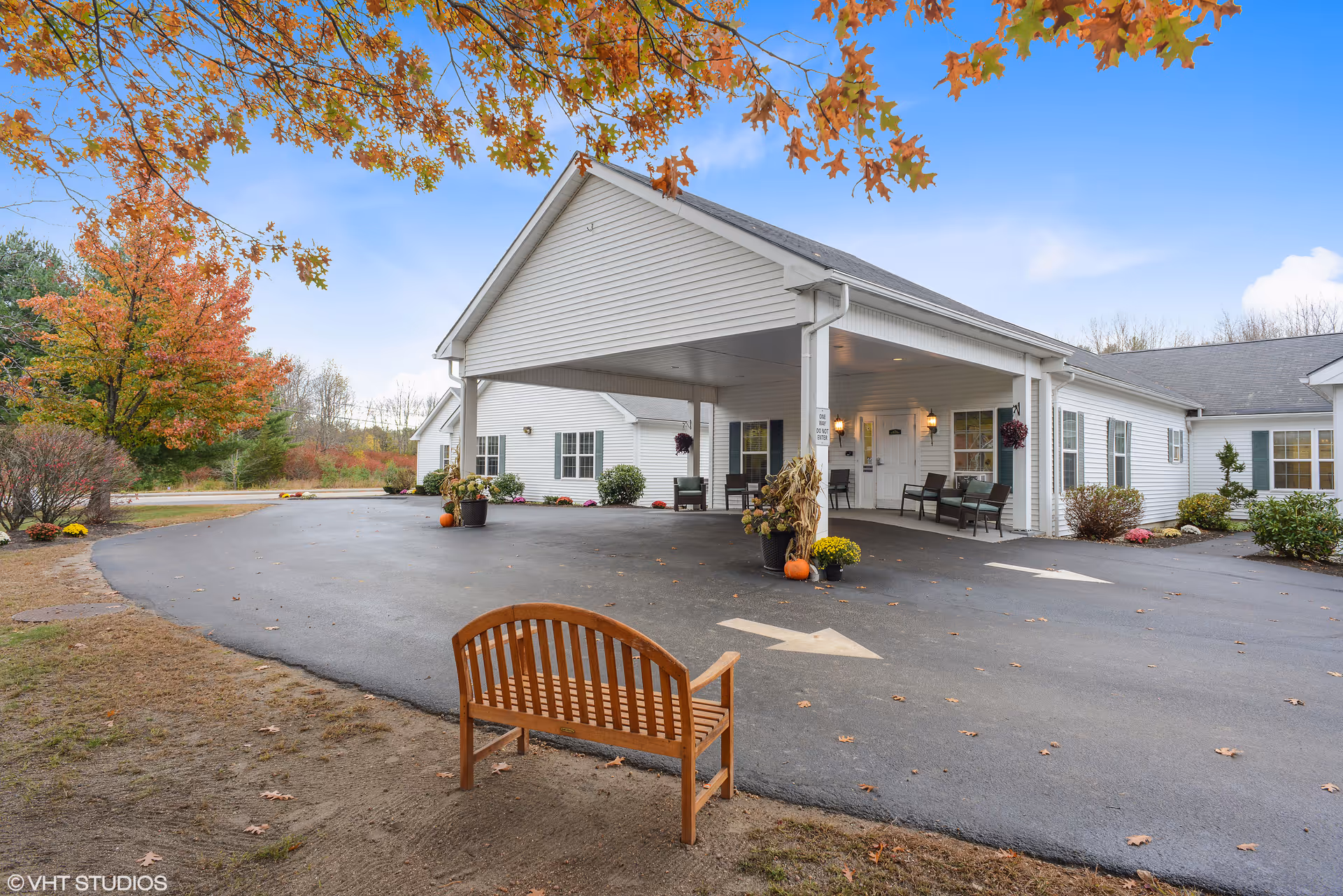 Exterior view of a single-story white building with a covered entrance driveway. There are benches and potted plants with autumn decorations near the entrance. Trees with fall foliage surround the area under a clear blue sky.