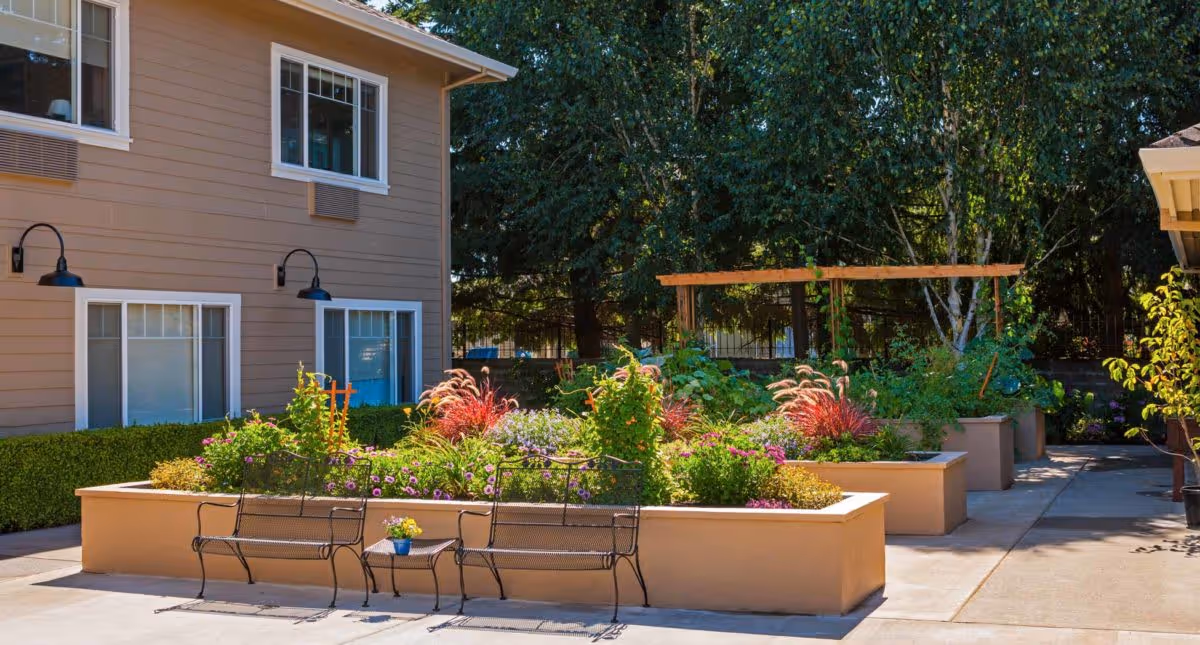 Outdoor garden area at The Hampton and Ashley Inn Memory Care Community featuring raised flower beds with colorful plants and flowers, two black metal benches with a small table in between, and a building with windows and wall-mounted lights in the background.