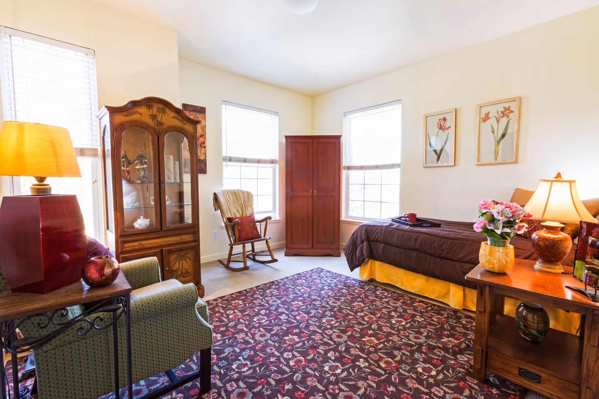 A cozy, well-lit bedroom with a single bed covered in a brown comforter and yellow bed skirt. The room features a wooden rocking chair, a wooden armoire, a green upholstered armchair, and two side tables with lamps and decorative items. The walls are adorned with two floral paintings, and the floor has a patterned area rug with floral designs. Large windows with blinds allow natural light to fill the room.