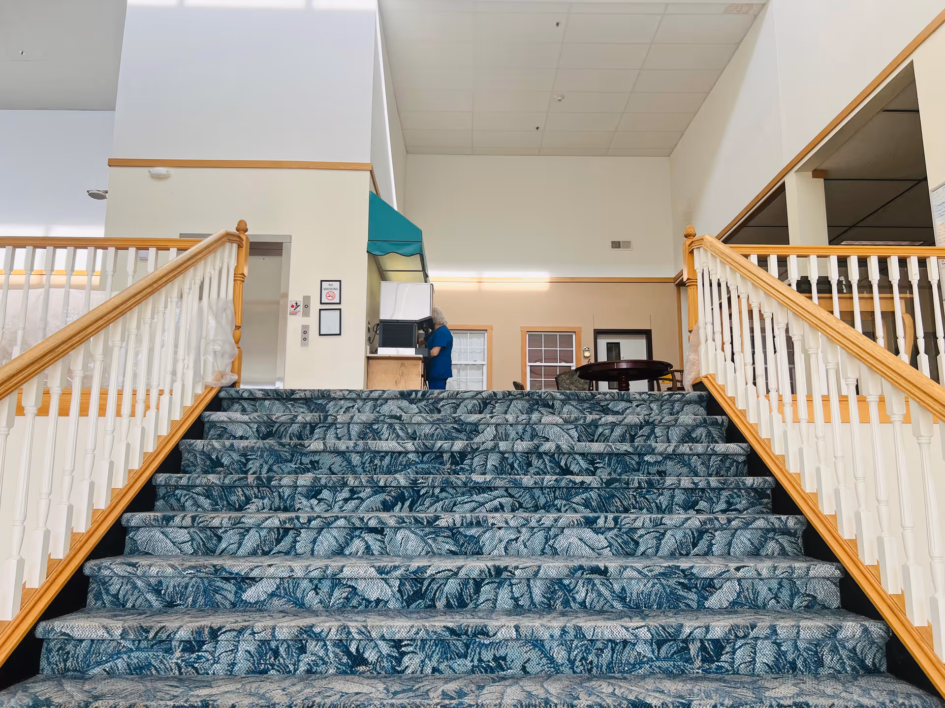 Wide carpeted staircase with a blue and green leaf pattern leading up to a landing area with wooden railings on both sides. At the top of the stairs, a person in blue scrubs is standing near a counter with a green awning above it. The walls are light-colored with a high ceiling and fluorescent lighting.