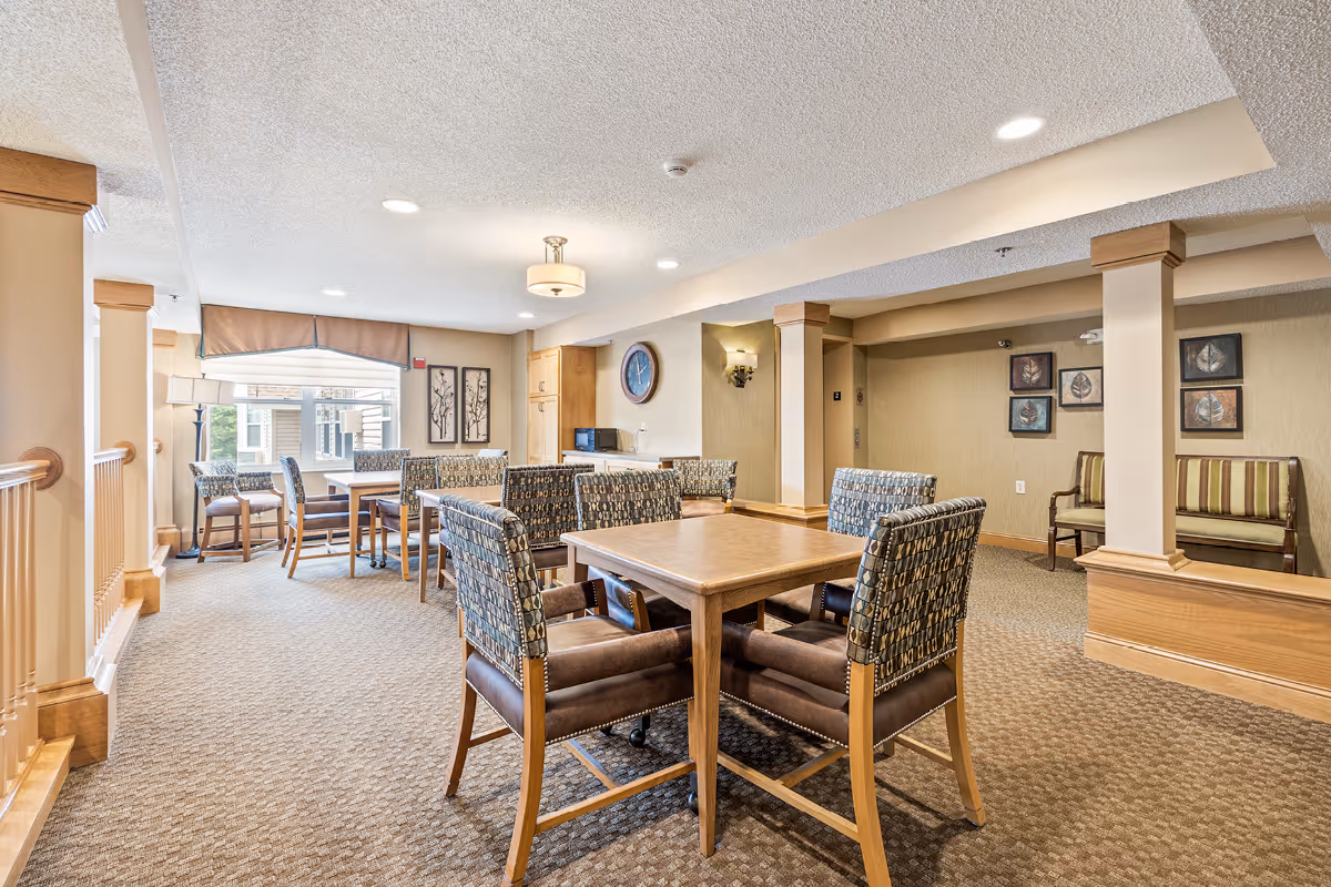 A well-lit common area in a senior living facility featuring multiple tables and cushioned chairs arranged for socializing or activities. The room has beige walls, carpeted floors, decorative wall art, and a window with a valance allowing natural light to enter. There is a clock on the wall and a small kitchenette area with a microwave in the background.
