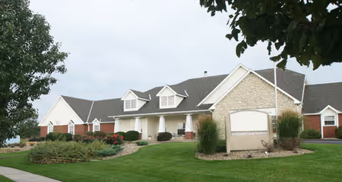Exterior view of a single-story senior living facility building with a combination of brick and stone facade, a well-maintained lawn, shrubs, and a sign in front of the building. The sky is overcast and there are trees partially framing the image.