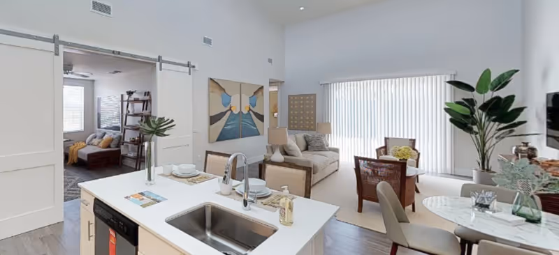 A bright and modern open-concept living space featuring a kitchen island with a sink and place settings, a cozy living room with a sofa and armchairs, a dining area with a round table and chairs, and a bedroom visible through a sliding barn door. The room is decorated with plants, artwork, and has large vertical blinds covering a window or sliding door.