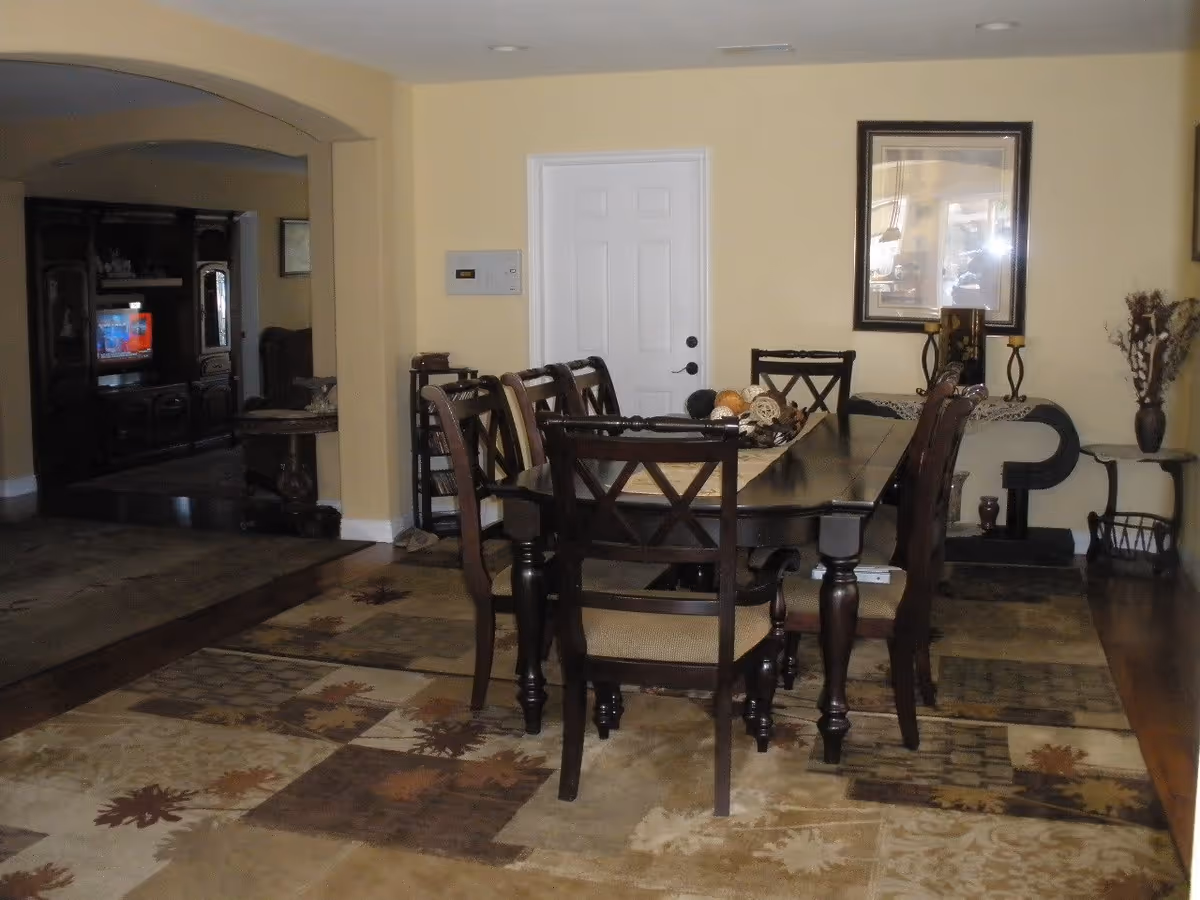 Dining room with a dark wooden dining table and six matching chairs on a patterned area rug. The walls are painted yellow, and there is a white door in the background. A framed picture hangs on the wall above a decorative console table with candles and a vase with dried flowers. An archway leads to a living room area with a TV and wooden entertainment center.
