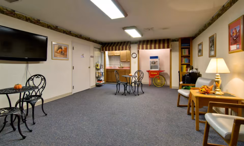 A senior living common room with small round tables and chairs, a popcorn machine by a kitchenette, a wall-mounted TV, and seating with side tables and a lamp.