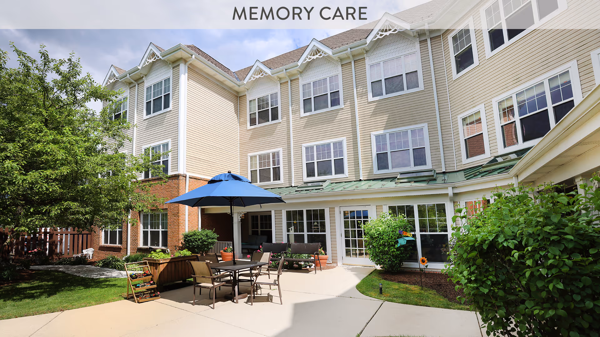 Outdoor patio area of a senior living facility with a table, chairs, and a blue umbrella. The building has beige siding with white trim and multiple windows. There are green bushes and trees surrounding the patio, and the words 'MEMORY CARE' are visible at the top of the image.