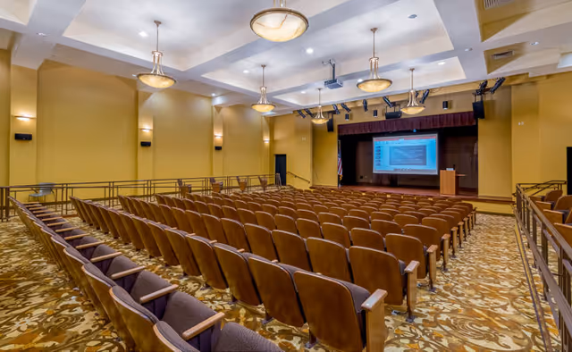 An auditorium with rows of brown cushioned seats facing a stage with a podium and a large screen displaying a presentation. The room has yellow walls, patterned carpet, and ceiling lights.