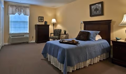 A cozy bedroom with a single bed covered in a blue quilt and white bed skirt, a dark wooden headboard, and a tray with a cup and plate on the bed. The room has beige walls, a window with blinds and a valance, a dark wooden dresser, a patterned armchair with a small side table and lamp, and a nightstand with a lamp and decorative items.
