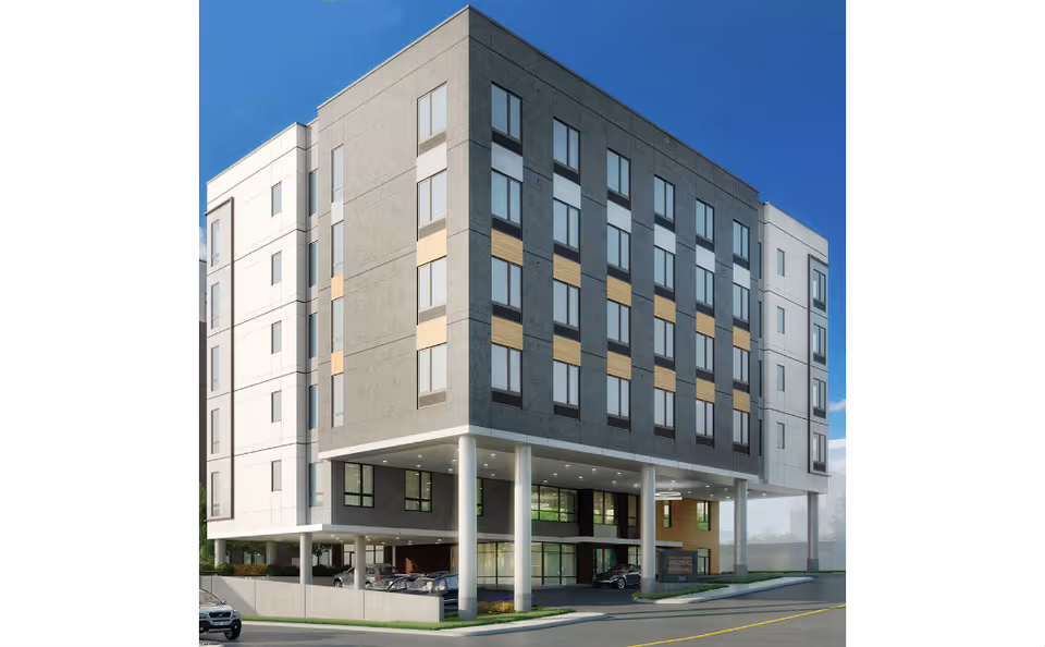 Exterior view of a modern multi-story senior living facility building with large windows, supported by white pillars, and a covered parking area underneath. The sky is clear and blue.