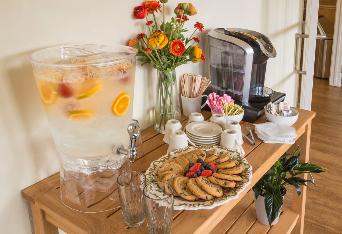 A wooden table set up with a large clear beverage dispenser filled with ice water and slices of lemon and orange, a plate of chocolate chip cookies garnished with berries, a coffee maker, cups, plates, napkins, sugar packets, stir sticks, and a vase of colorful flowers.