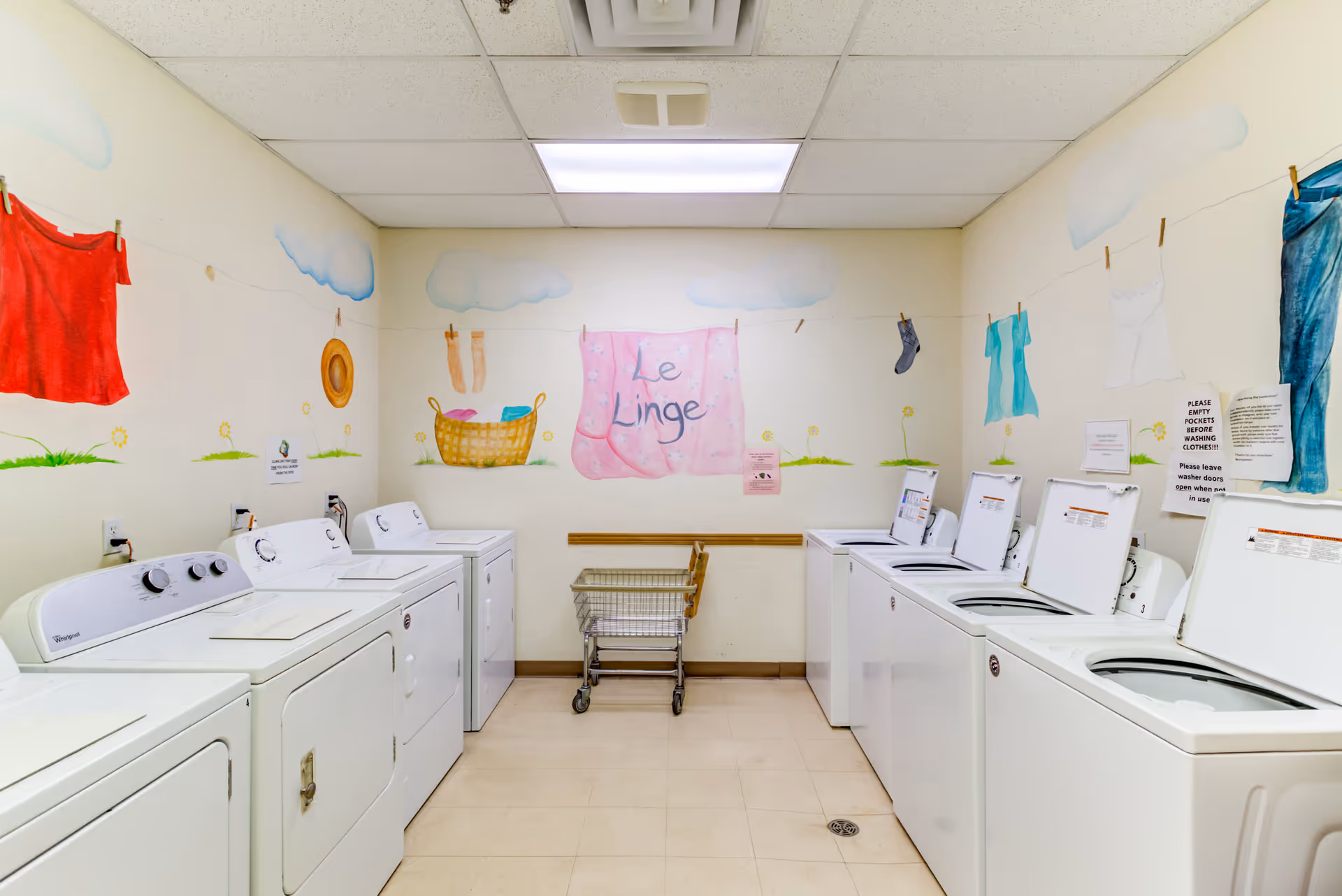 Laundry room with multiple white washing machines and dryers along both walls. The walls are decorated with colorful murals of clothes hanging on a clothesline, clouds, grass, and flowers. A laundry cart is positioned against the back wall beneath a mural that reads 'Le Linge'.