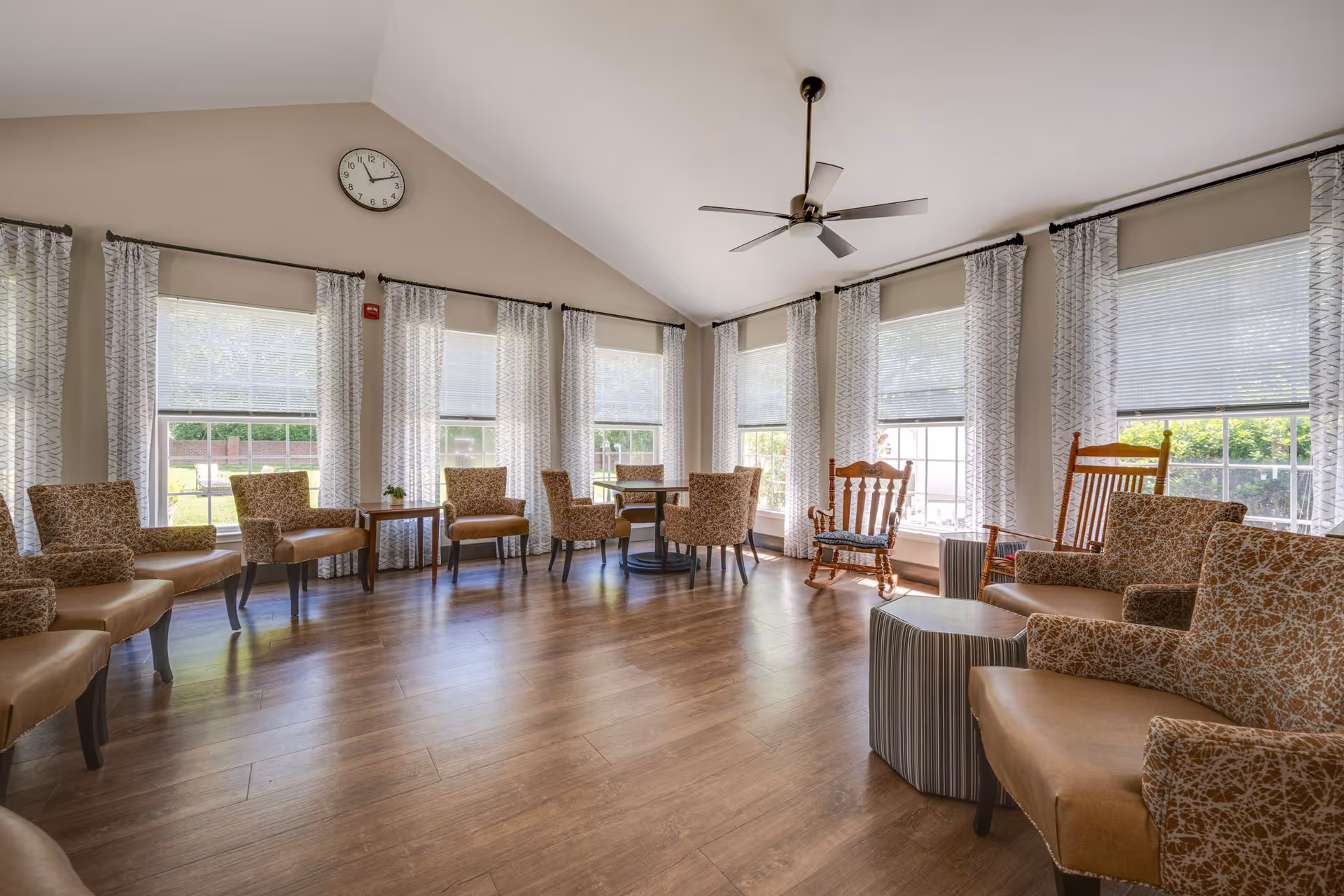 A bright and spacious living room with multiple patterned armchairs arranged around small tables. The room features large windows with white blinds and patterned curtains, a ceiling fan, and a wall clock above the windows. The floor is wooden, and the room has a high vaulted ceiling.
