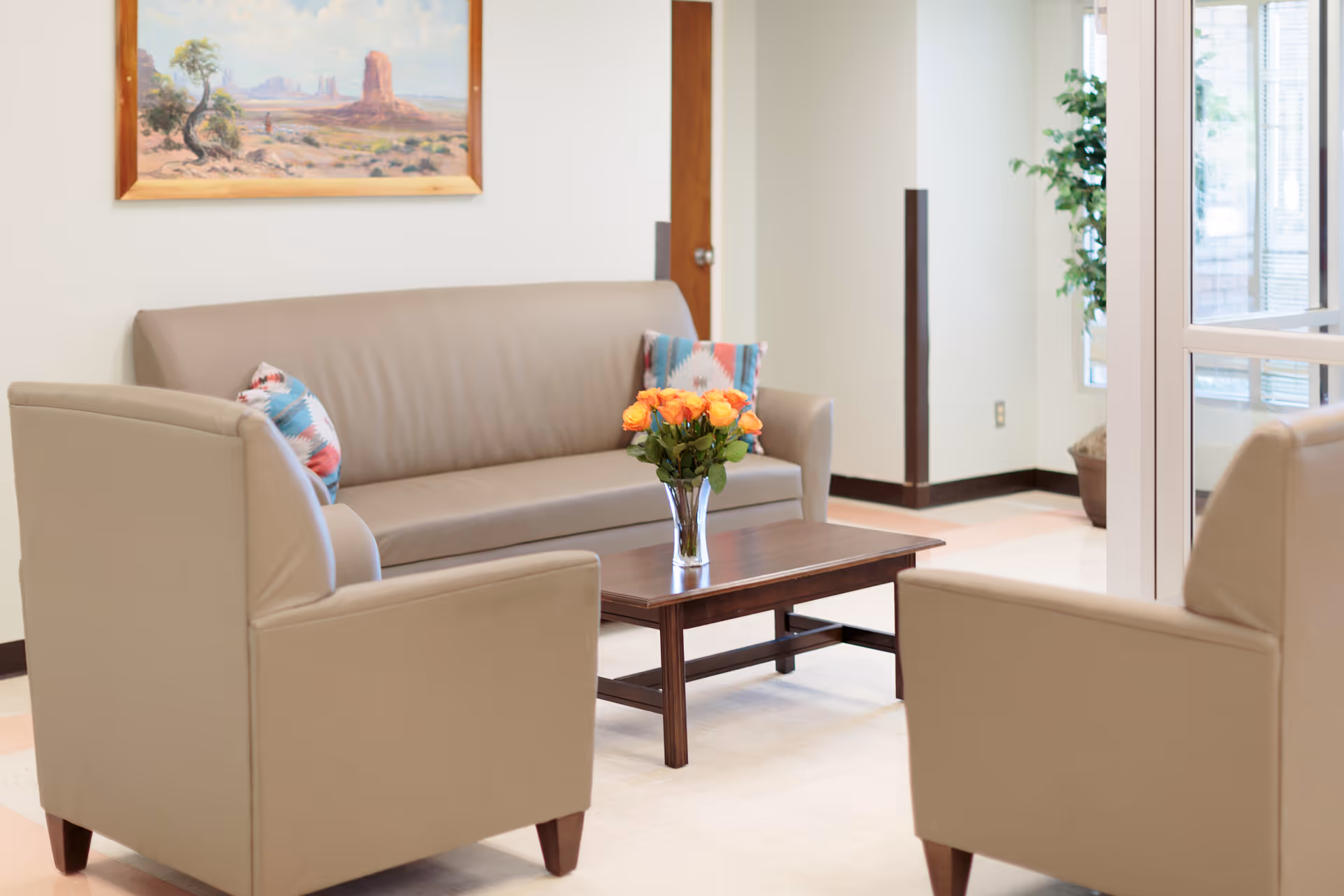 A seating area with a beige leather sofa and two matching armchairs arranged around a wooden coffee table with a vase of orange roses. There are two patterned cushions on the sofa, a framed landscape painting on the wall, and a potted plant near a glass door in the background.