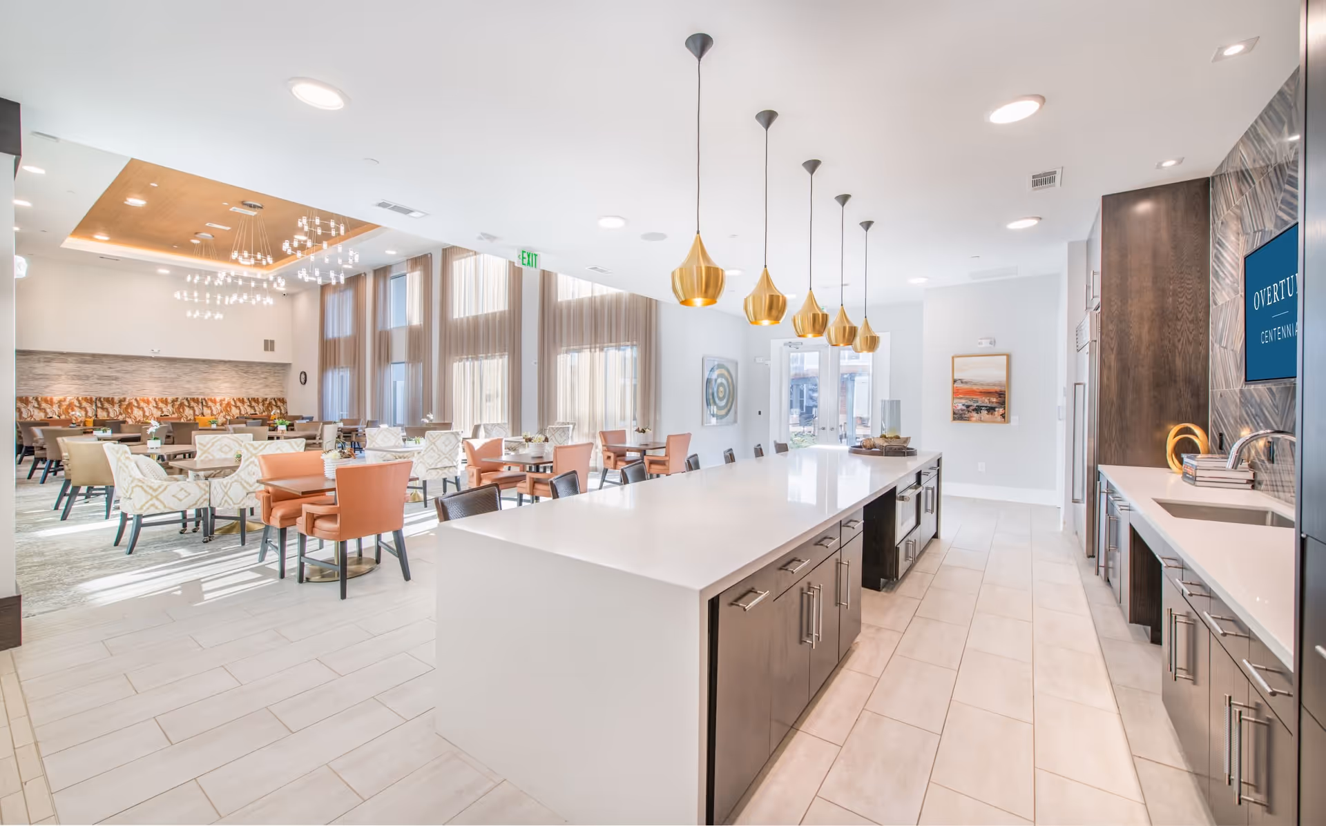 A bright, modern dining and kitchen area in a senior living facility. The kitchen features a long white island with dark cabinets and six gold pendant lights hanging above. The dining area has multiple tables with a mix of beige and orange chairs, large windows with sheer curtains, and contemporary light fixtures on the ceiling.