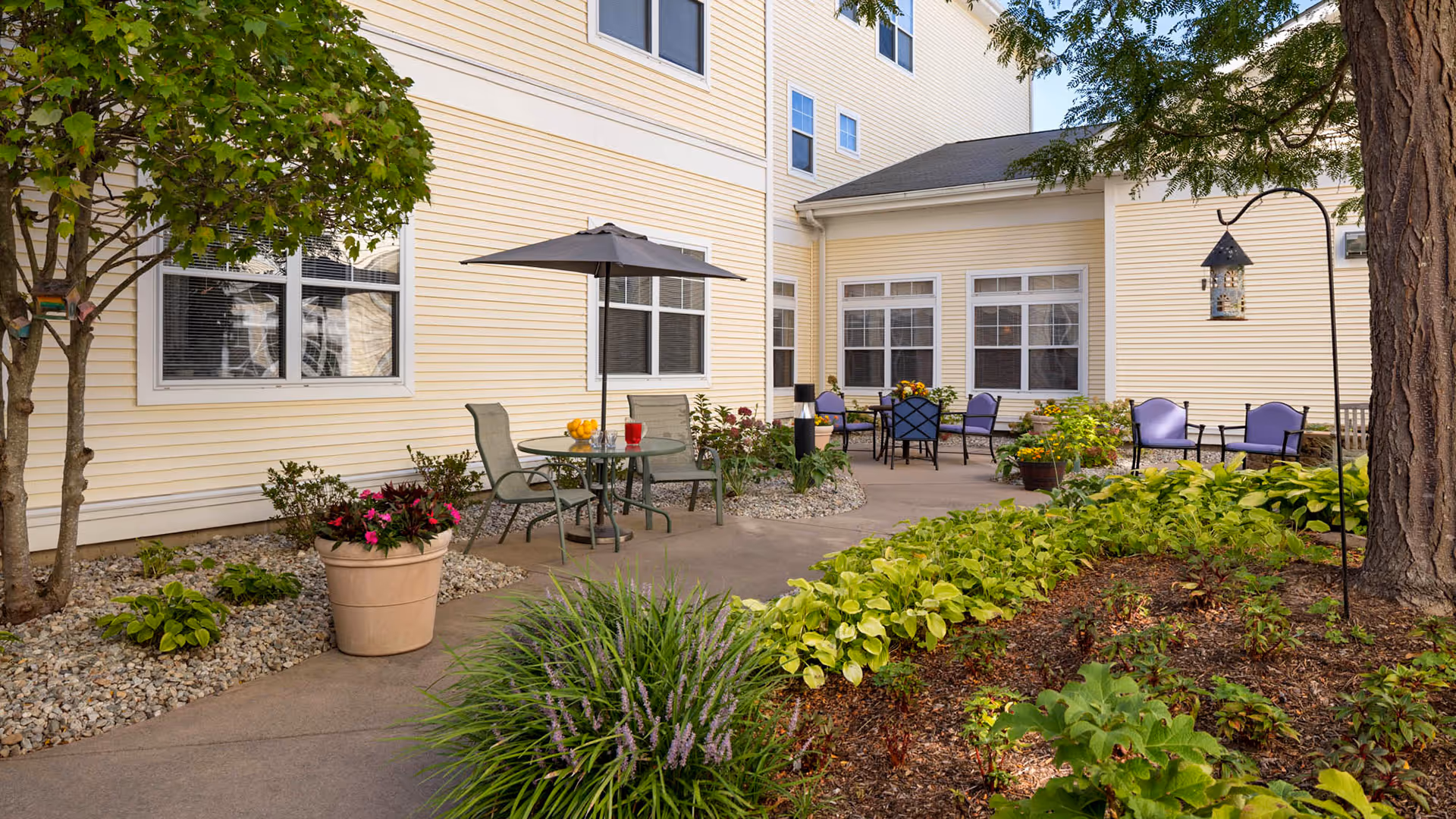Outdoor courtyard patio with tables, chairs, an umbrella, and landscaped plantings beside a light-yellow building.