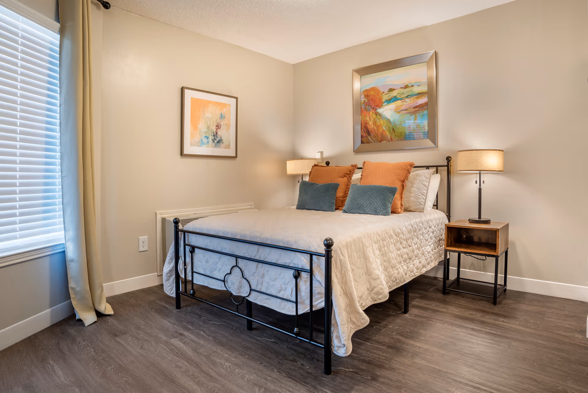 A bedroom with a black metal bed frame, a white quilted bedspread, two orange pillows and two blue pillows. There is a wooden nightstand with a lamp on the right side of the bed. The walls are beige with two framed paintings, one above the bed and one on the adjacent wall. A window with white blinds and beige curtains is on the left side of the room. The floor is a dark wood laminate.